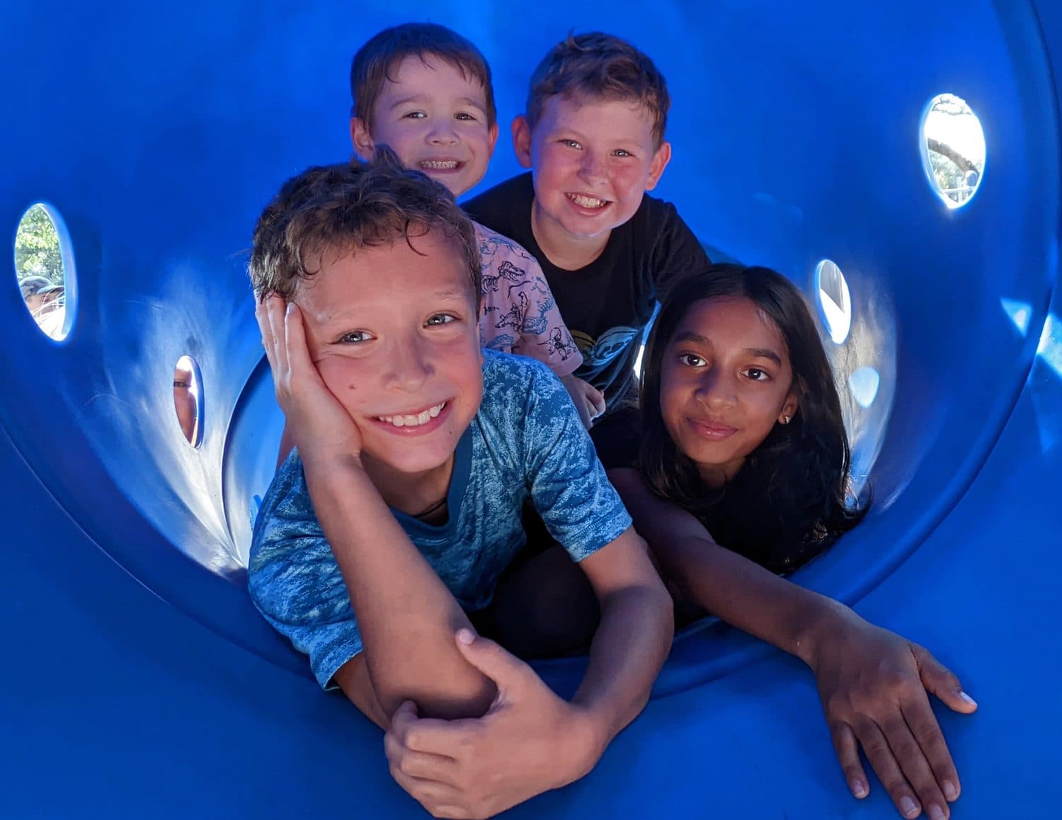 Four Crystal Challenges students packed into a blue tunnel slide at recess, grinning at the camera.